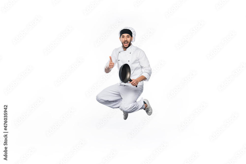joyful chef in hat and uniform jumping, showing thumb up while holding frying pan isolated on white.