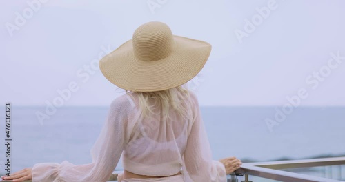 Stunning blonde woman in summer beach outfit relaxing outdoors against sea background. Back view of fashionable romantic young adult lady wearing a trendy vintage straw hat, white blouse, and skirt photo