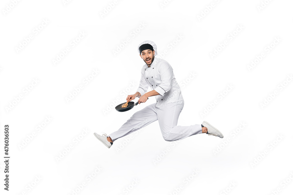 happy chef in hat and uniform jumping and holding frying pan with wooden spatula isolated on white.