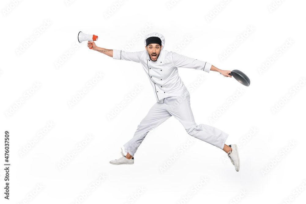 surprised young chef in hat and uniform levitating while holding frying pan and megaphone isolated on white. surprised young chef in hat and uniform levitating while holding frying pan and megaphone isolated on white.