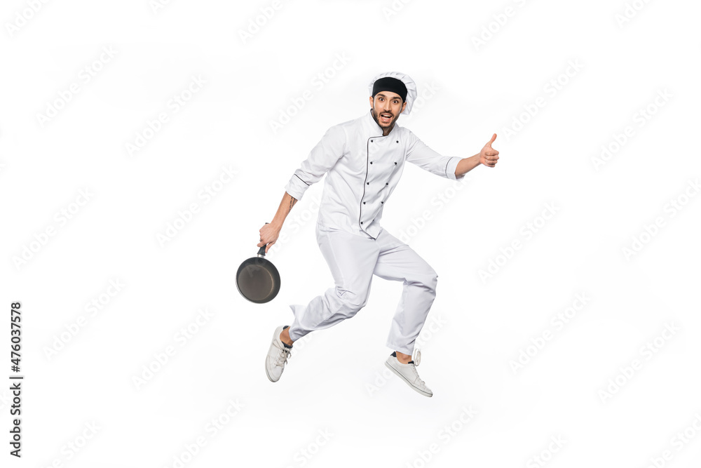 cheerful chef in hat and uniform jumping, showing thumb up while holding frying pan isolated on white. cheerful chef in hat and uniform jumping, showing thumb up while holding frying pan isolated on white.