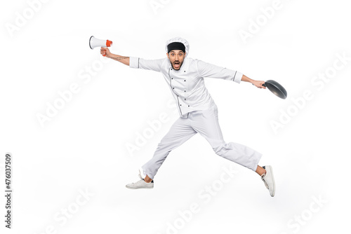 surprised young chef in hat and uniform levitating while holding frying pan and megaphone isolated on white. © LIGHTFIELD STUDIOS