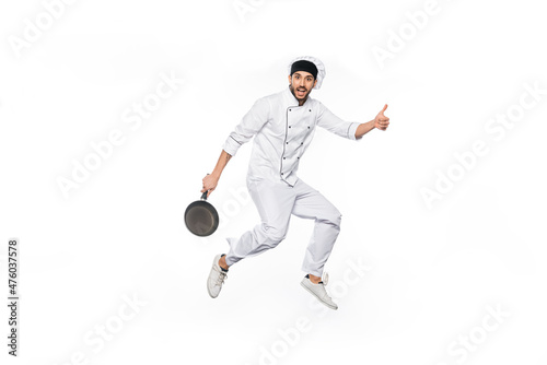 cheerful chef in hat and uniform jumping, showing thumb up while holding frying pan isolated on white.
