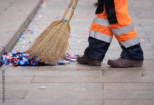 A janitor with a broomstick removes bright colorful confetti from the side of a city street after a celebration event
