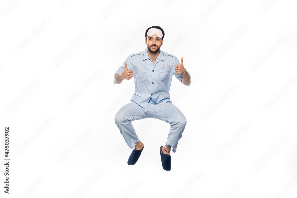 happy bearded man in pajamas and sleeping mask levitating while showing thumbs up on white.