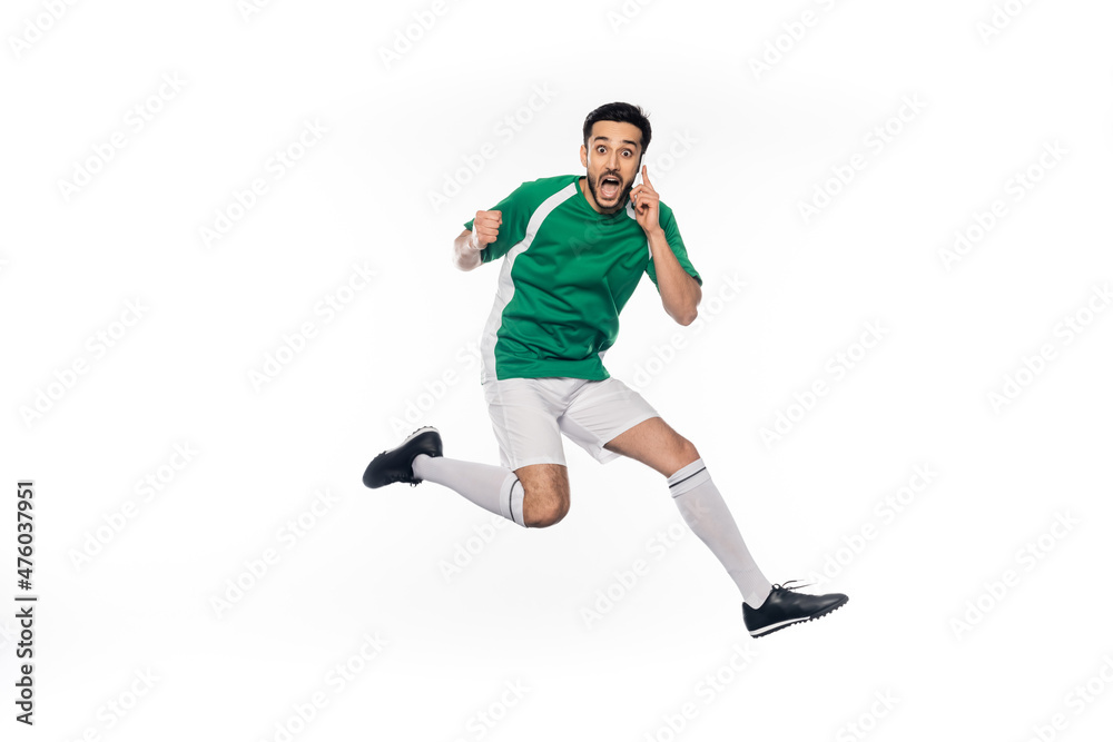 amazed football player in uniform jumping while talking on smartphone on white.