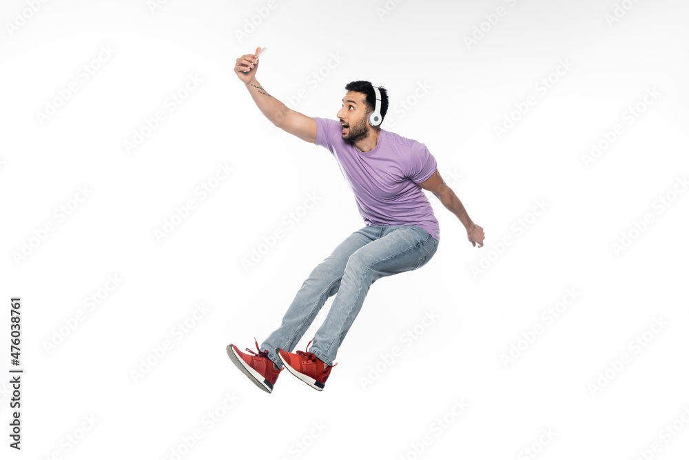 cheerful man in wireless headphones taking selfie and levitating on white.