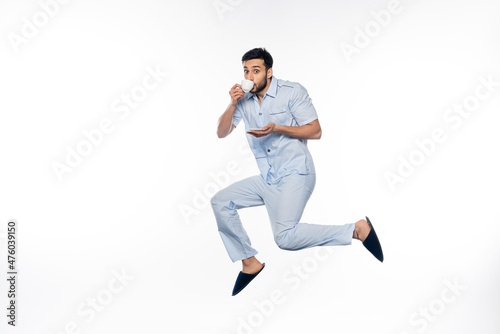 bearded man in pajamas holding cup and drinking coffee while levitating on white.