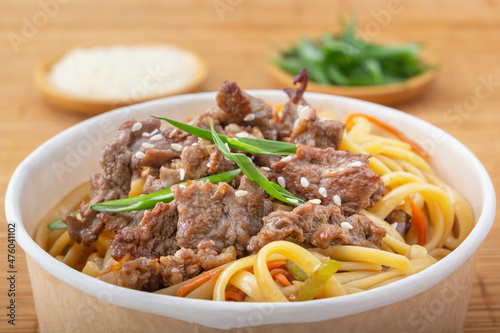 Noodles with meat, pepper, herbs and sesame seeds in a paper disposable plate on the background of a mat . Asian street food. Close-up