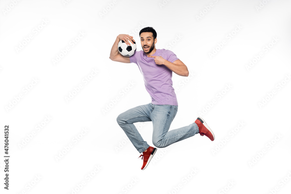 happy man in jeans and purple t-shirt levitating while pointing at football on white.