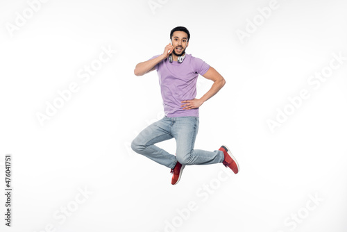 positive man in wireless headphones talking on cellphone and levitating on white.