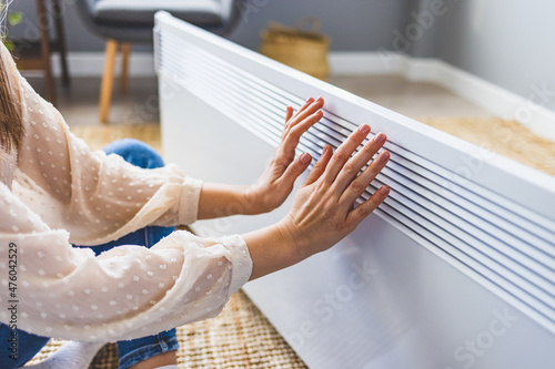 Woman warming hands near electric heater at home. Using heater at home in winter. Woman warming her hands. Heating season. Young woman warming hands near electric heater at home