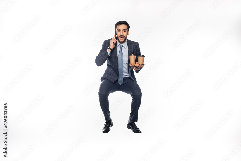 Obraz premium happy businessman in suit jumping while holding cup holder with paper cups and talking on smartphone on white.