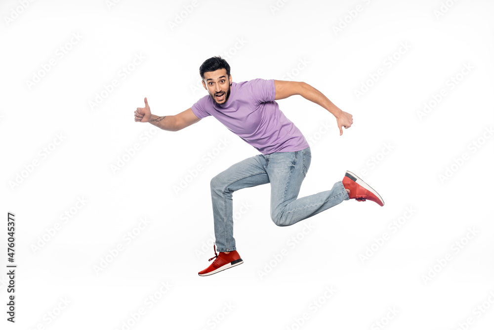 amazed man in jeans and purple t-shirt levitating while showing thumbs up isolated on white.