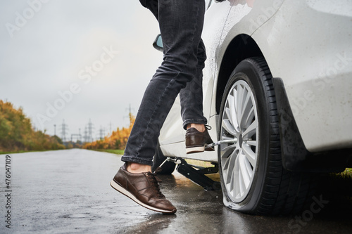 Close up of male auto mechanic standing on metal wrench while unscrewing lug nuts on car wheel. Young man using lug nut wrench while changing flat tire on the road. Concept of emergency road service. © anatoliy_gleb