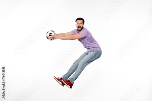 excited man in jeans and purple t-shirt levitating with football on white.