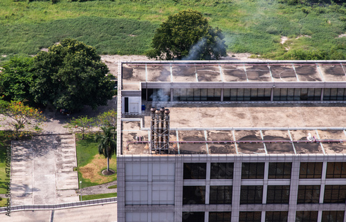 Smoke coming out of the four metal Chimney on the flat roof off a big building in the city. No focus, specifically.