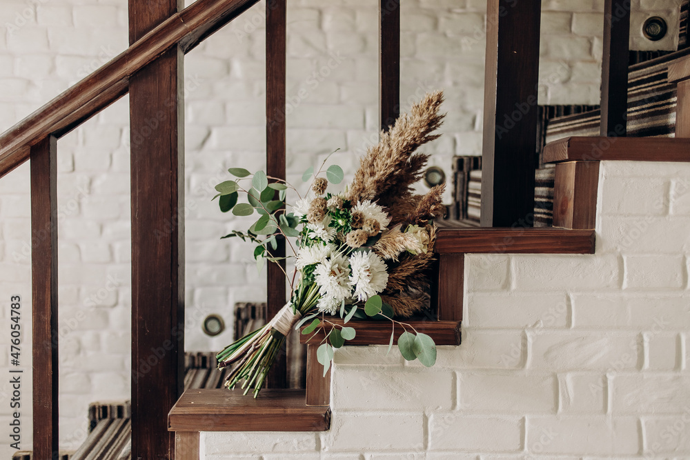A beautiful boho style bridal bouquet is attached to a wooden railing, which is part of the staircase, inside the house. A wedding bouquet in a boho style lies against the background of a white brick 