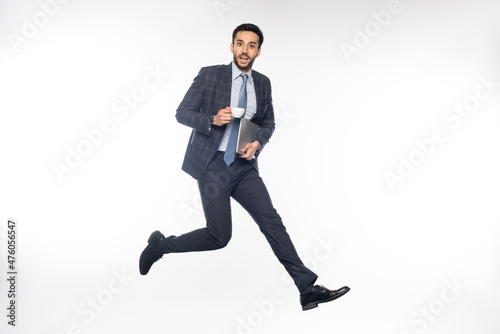 excited businessman in suit jumping while holding laptop and cup on white.