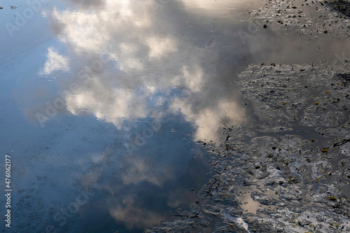 Blue cloudy sky reflected in the water of a ditch with oily substance on the surface and caked gunk on the right. Pollution photo