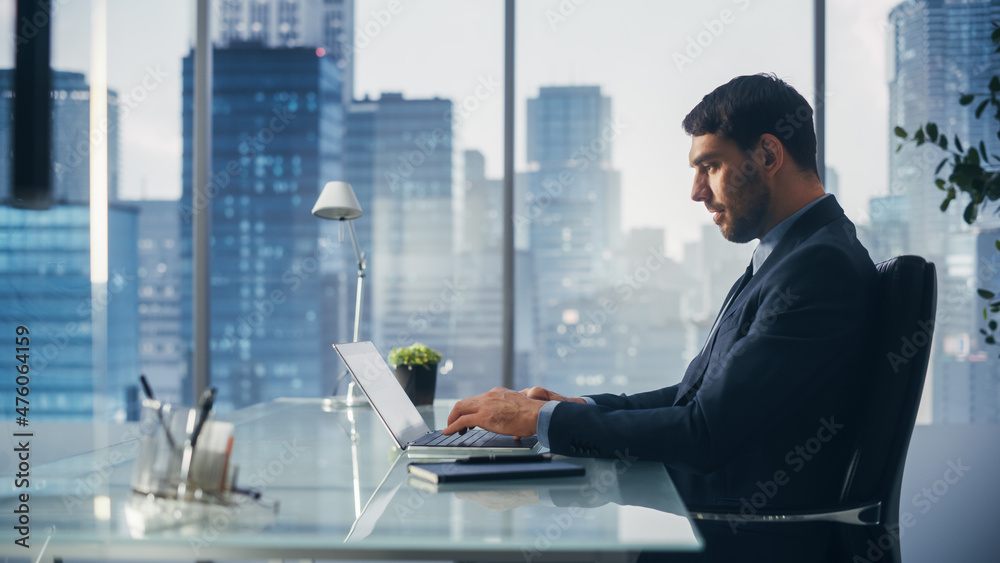 Confident Businessman in a Suit Sitting at a Desk in Modern Office, Using Laptop Computer, Next to Window with Big City with Skyscrapers View. Successful Finance Manager Planning Work Projects. Confident Businessman in a Suit Sitting at a Desk in Modern Office, Using Laptop Computer, Next to Window with Big City with Skyscrapers View. Successful Finance Manager Planning Work Projects.