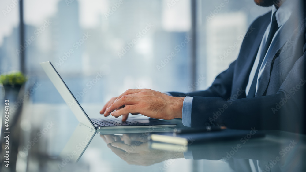 Close Up on Businessman Sitting at a Desk in Modern Office, Using Laptop Computer, Next to Window with Big City with Skyscrapers View. Successful Finance Manager Planning Work Projects. Close Up on Businessman Sitting at a Desk in Modern Office, Using Laptop Computer, Next to Window with Big City with Skyscrapers View. Successful Finance Manager Planning Work Projects.