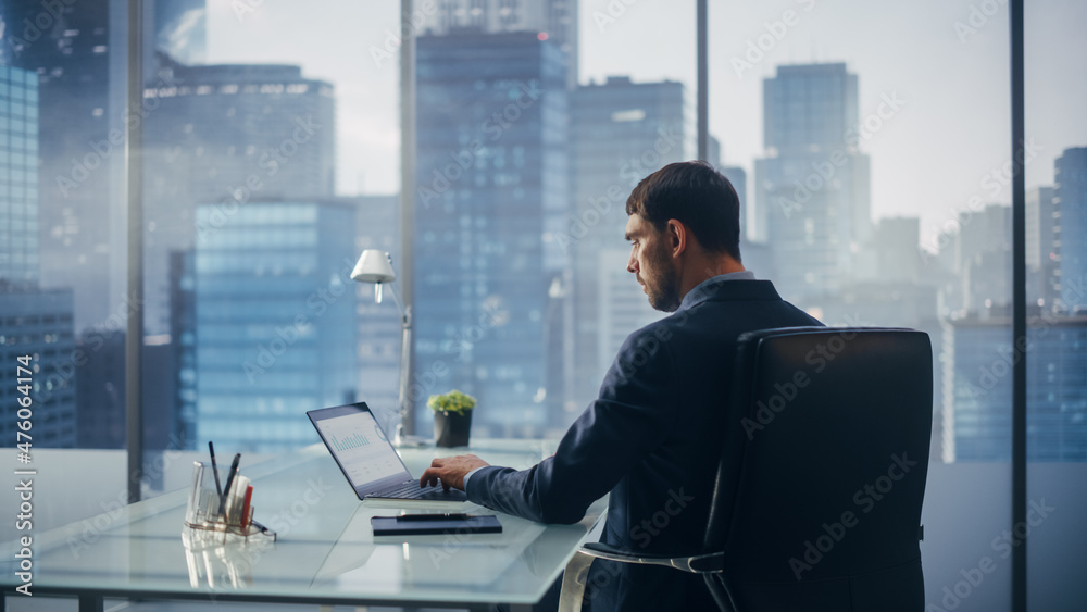 Relaxed Businessman in a Suit Sitting at a Desk in Modern Office, Using Laptop Computer, Next to Window with Big City with Skyscrapers View. Successful Finance Manager Planning Work Projects. Relaxed Businessman in a Suit Sitting at a Desk in Modern Office, Using Laptop Computer, Next to Window with Big City with Skyscrapers View. Successful Finance Manager Planning Work Projects.