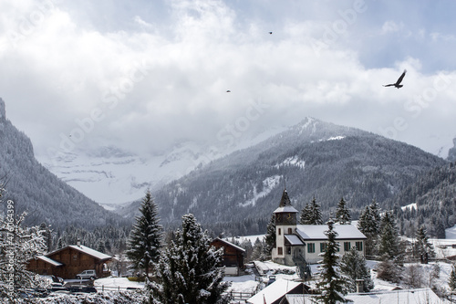 Village scenery with the church in the middle with flying birds in the blue and cloudy sky and with mountains in the background in the winter, Switzerland