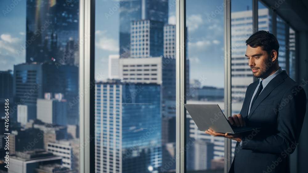 Businessman is Standing in Modern Office, Using Laptop Computer, Next to Window with Big City with Skyscrapers View. Successful Corporate CEO or Finance Manager Planning Investment Work Projects. Businessman is Standing in Modern Office, Using Laptop Computer, Next to Window with Big City with Skyscrapers View. Successful Corporate CEO or Finance Manager Planning Investment Work Projects.