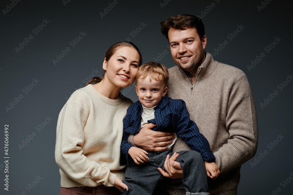 Waist up portrait of adult couple holding cute son and looking at camera in studio against black backckground Waist up portrait of adult couple holding cute son and looking at camera in studio against black backckground
