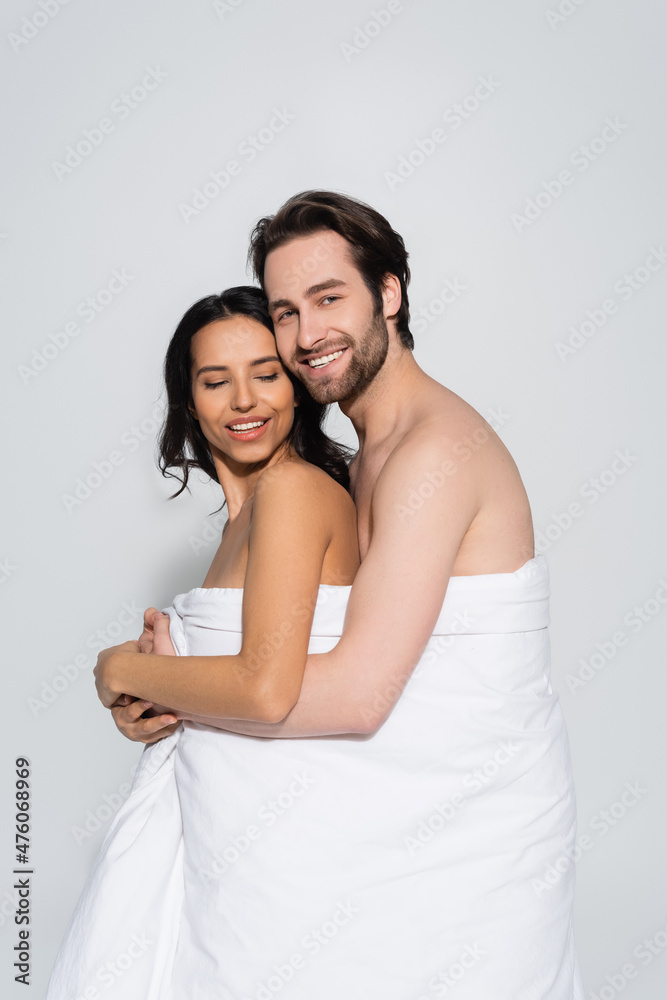 cheerful man looking at camera while hugging sexy woman in white blanket isolated on grey. cheerful man looking at camera while hugging sexy woman in white blanket isolated on grey.