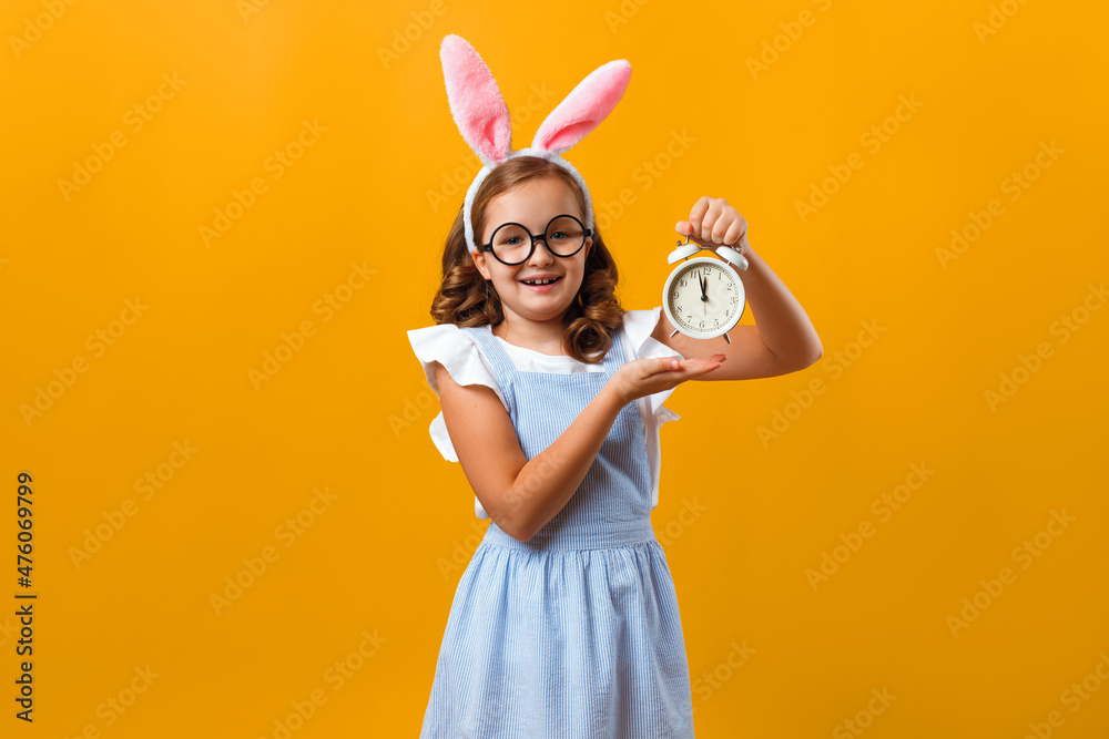 Happy little girl in Easter bunny ears on isolated yellow background. The child is holding an alarm clock.