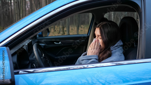 Woman driver having cold and with runny nose coughs and blows nose into paper napkin sitting at steering wheel of car on roadside © lenblr