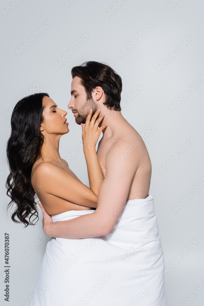 side view of shirtless man and passionate woman in white blanket embracing while standing face to face isolated on grey. side view of shirtless man and passionate woman in white blanket embracing while standing face to face isolated on grey.