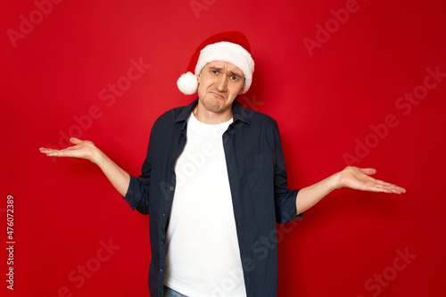 Horizontal shot of attractive man with puzzled expression spreads hands with confused look, wears christmas hat feels hesitant indignant isolated over red background. concept - doubt, new year, people