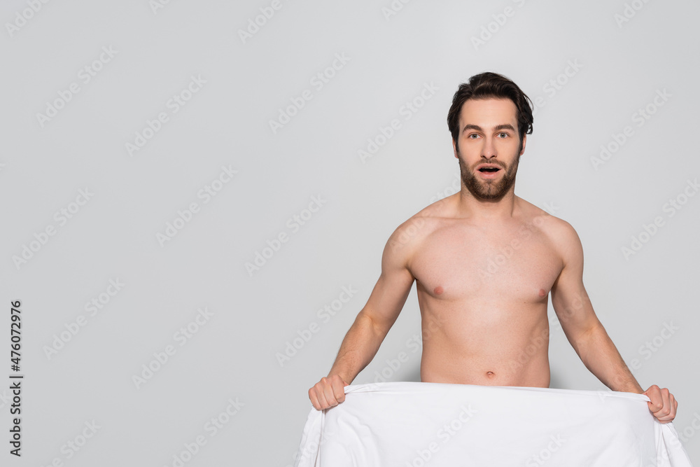 Fototapeta premium astonished shirtless man holding white blanket while looking at camera on grey.