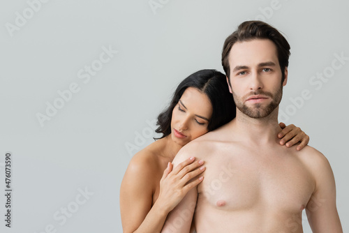 sexy shirtless man looking at camera near seductive brunette woman hugging him isolated on grey.