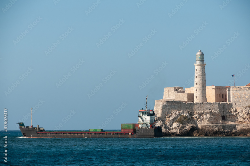 Medium container boat leaving the bay of Havana with the Morro Castle lighthouse on the shore.