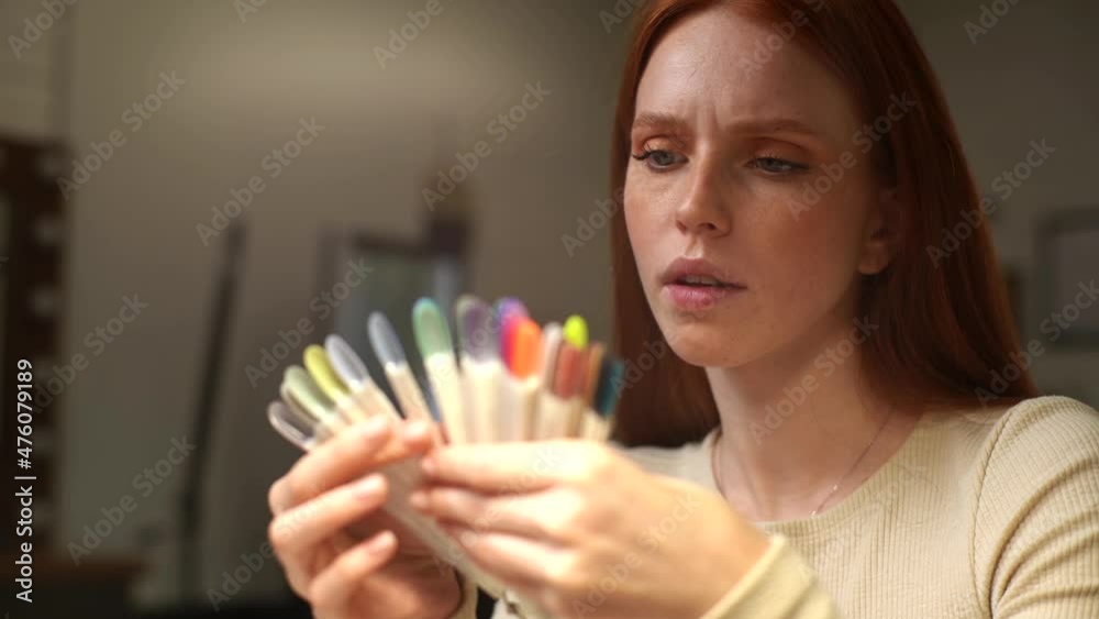 Close-up face of focused pretty young woman client choosing colour from colourful nails tips in manicure salon. Thoughtful redhead female choosing perfect nail varnish color. Shooting in slow motion. Close-up face of focused pretty young woman client choosing colour from colourful nails tips in manicure salon. Thoughtful redhead female choosing perfect nail varnish color. Shooting in slow motion.