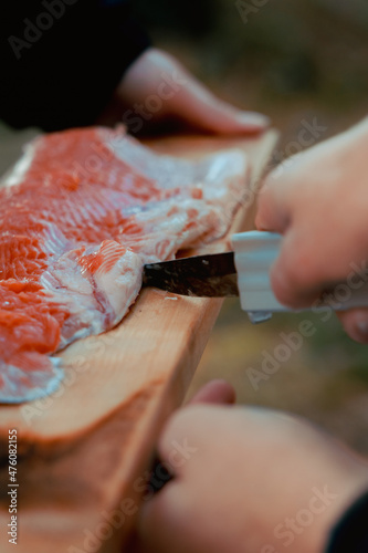 Young people making a salmon by campfire for the first time. Hands cutting the fish with knife. Wooden cutting board under the food.