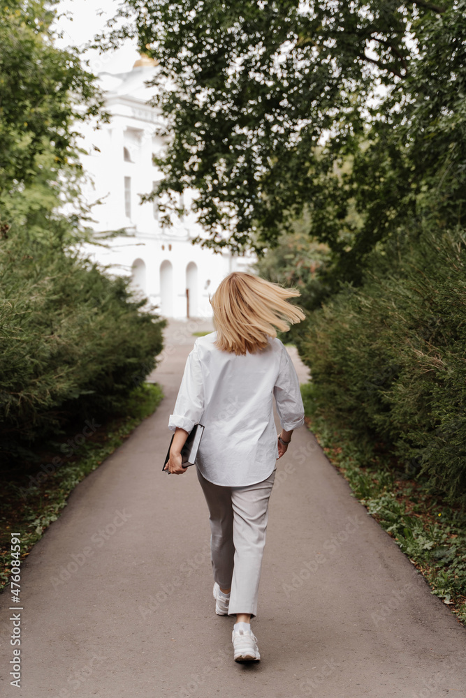 blonde in white clothes walks through the park to the university with a book in her hands green plants trees white building. vertical blonde in white clothes walks through the park to the university with a book in her hands green plants trees white building. vertical