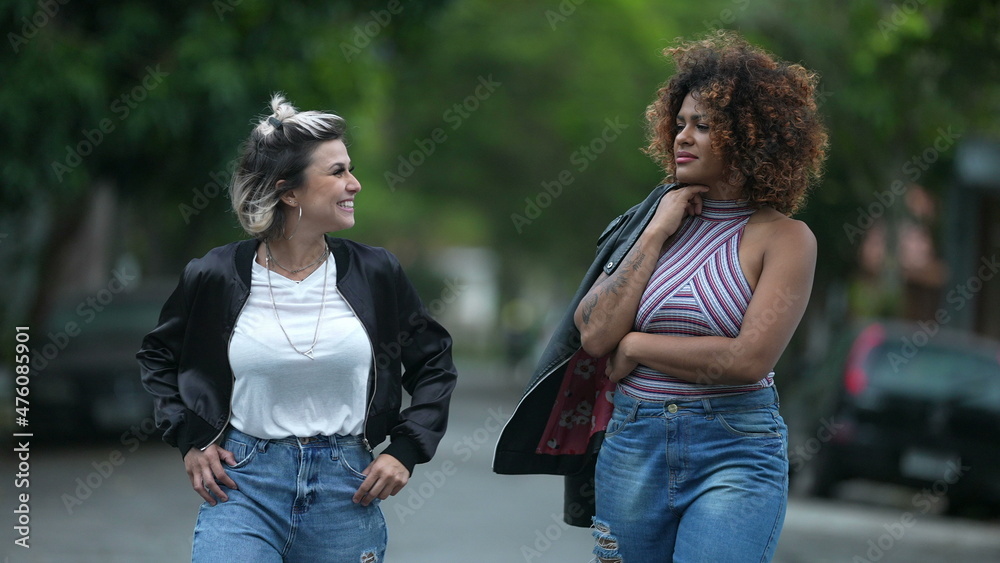 Fototapeta premium Candid two diverse women walking together outside in street laughing and smiling