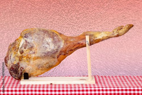 Spanish food. Closeup of a front leg of a Spanish Serrano ham on a wooden stand on a red checkered tablecloth over abstract pink cube block raster background. Template for your design.