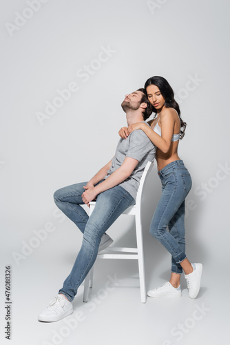 full length view of brunette woman in jeans and bra hugging man sitting on chair on grey.