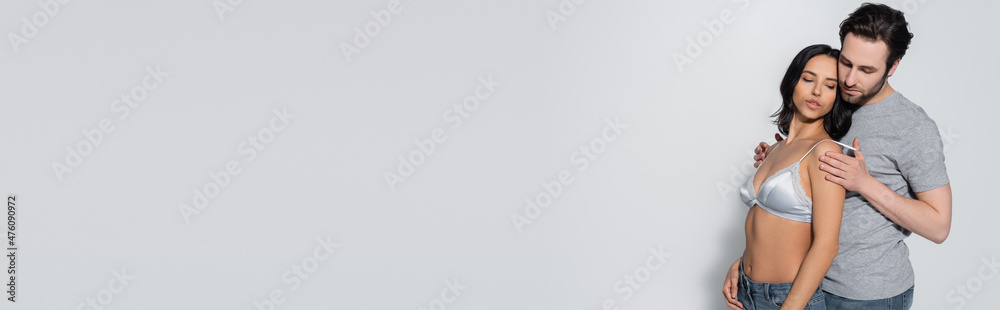 Naklejka premium bearded man in t-shirt hugging brunette woman in satin bra on grey, banner.