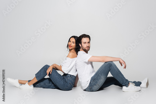 full length view of young couple in white t-shirts and jeans sitting back to back on grey.