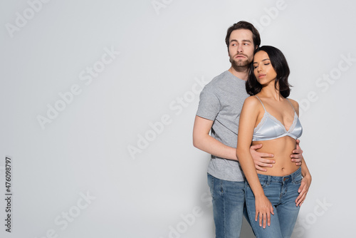 young man in t-shirt looking away while hugging hot woman in satin bra on grey.