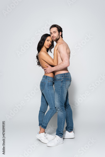 full length view of young couple in jeans and sneakers looking at camera while embracing on grey.