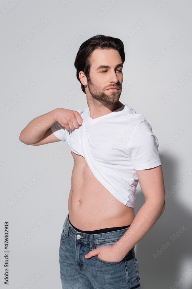 sexy man raising white t-shirt while standing with hand in pocket of jeans on grey. sexy man raising white t-shirt while standing with hand in pocket of jeans on grey.
