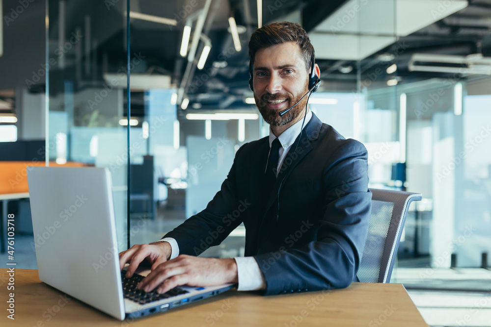 An employee of the call center looks at the camera and smiles uses a headset for a video dink, a businessman makes an online meeting with a laptop